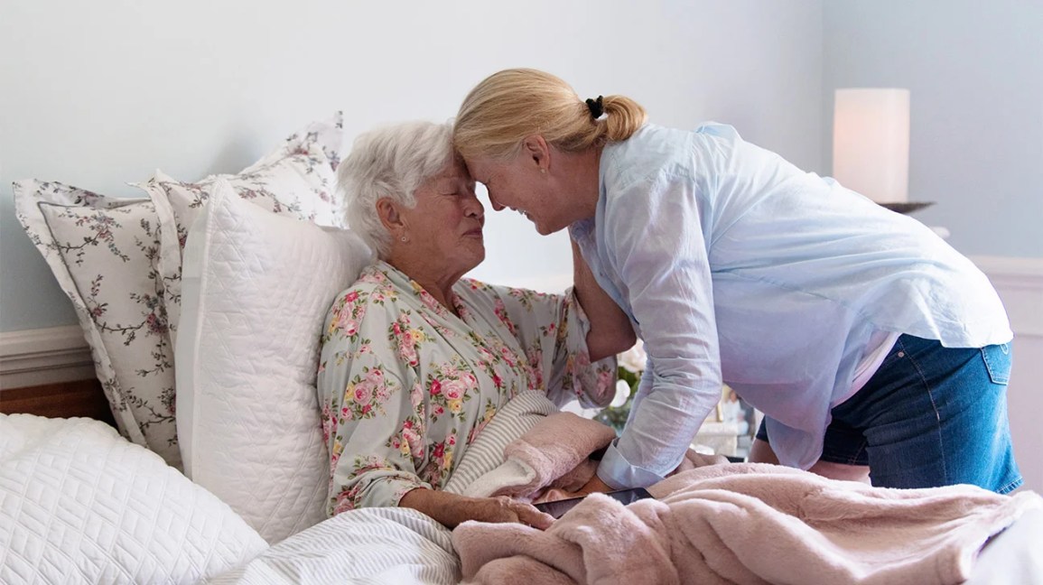 Older woman sharing an emotional moment with her mother, who is seated in a bed at home.