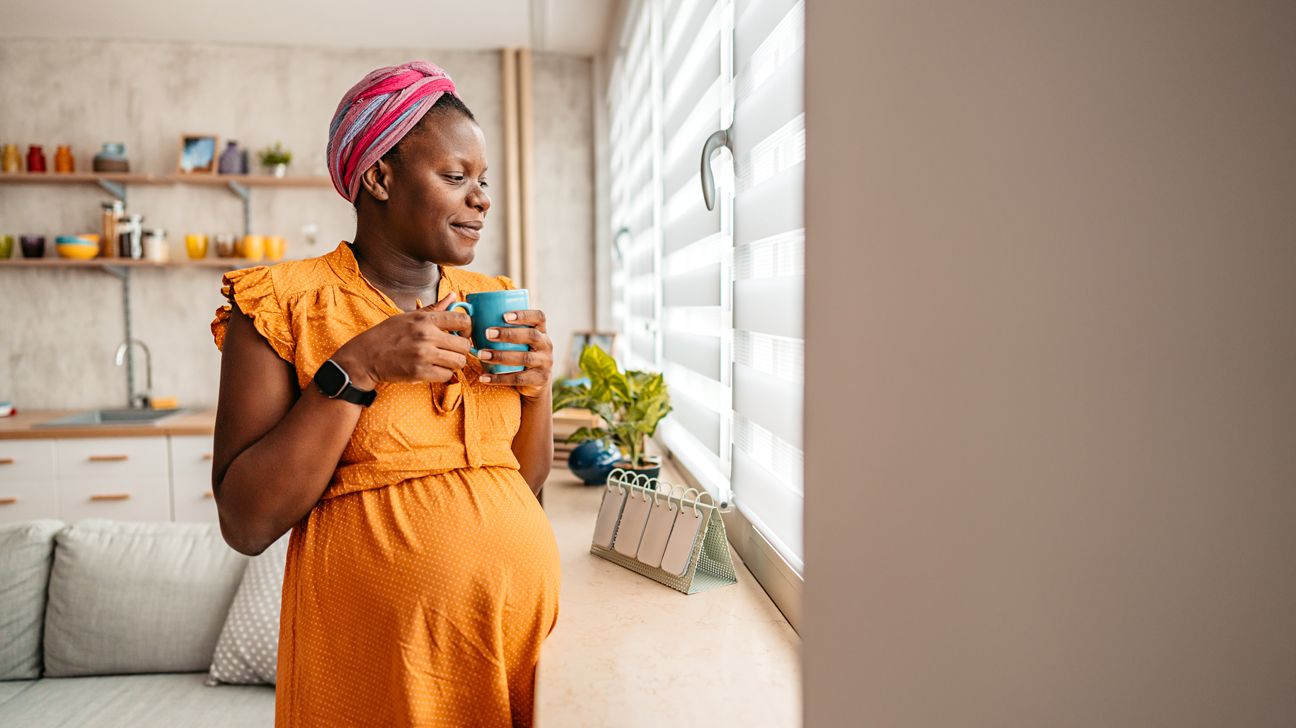 Pregnant woman standing by window drinking tea